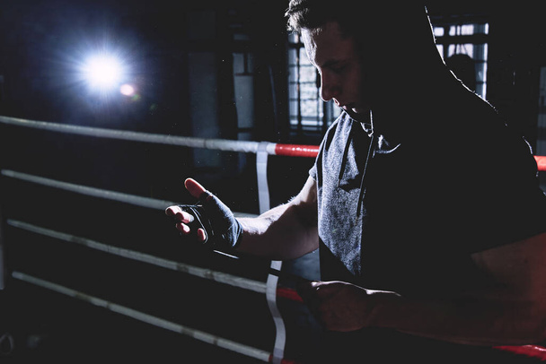 Primer plano retrato boxeador profesional en una capucha envuelve las palmas de sus manos, se pone guantes en el ring. Colores oscuros
 - Foto, imagen
