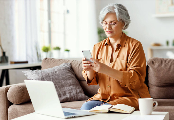 Старшая женщина сидит на диване рядом со столом с блокнотом и ноутбуком и просматривает смартфон во время работы над удаленным проектом на дому
 - Фото, изображение