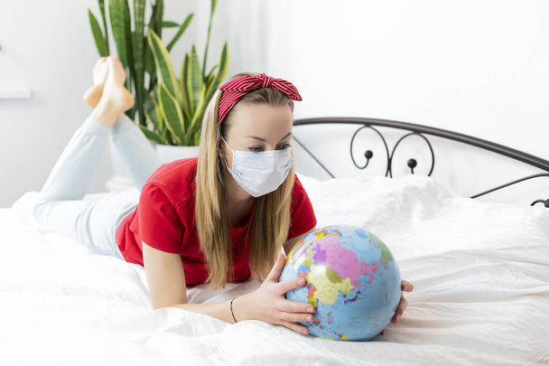 Tıbbi maskeli ve kırmızı tişörtlü genç hasta bir kadın beyaz bir yatakta uzanıyor ve dünyaya bakıyor. Seyahat edemez çünkü koronavirüs salgını nedeniyle uçuşlar iptal edildi. salgın COVID-19 - Fotoğraf, Görsel