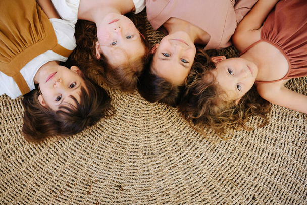 Quatro garotinhas giras da mesma idade estão deitadas no chão em um tapete de vime cabeça a cabeça. Todos olhando para a câmera
. - Foto, Imagem