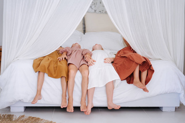 Four cute little girls about the same age are lying back on a double bed with curtains. All wearing dresses. Tiny bare feet hanging in the air. - Fotografie, Obrázek
