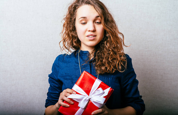 Curly girl holding a gift red box with white bow. Gray background. - Fotó, kép