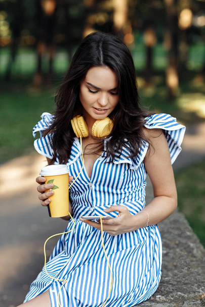 Young charming woman listening music with headphones while holding smartphone and coffee cup outdoor. - Foto, imagen