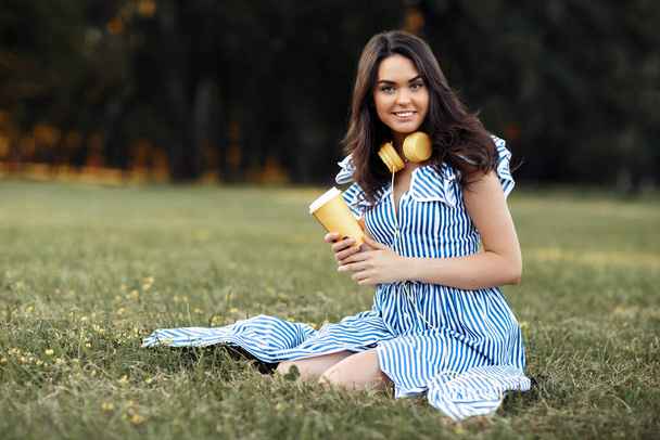 Happy young woman with headphones holding paper cup of tea or coffee at the park. - Foto, Bild