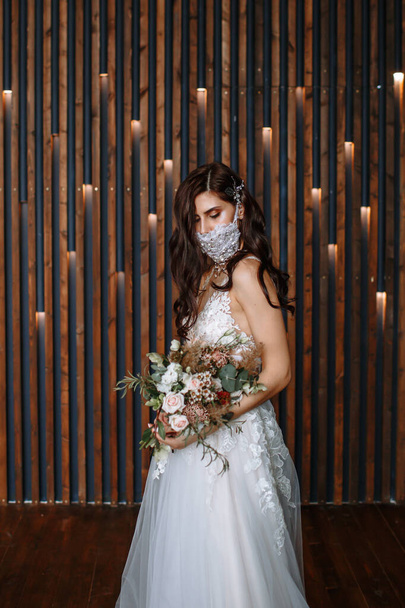 Невеста со свадебной противовирусной маской ручной работы на лице. Свадебный букет в руке
 - Фото, изображение