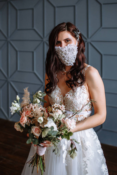 Bride in with a handmade wedding antiviral mask on her face. Wedding bouquet in hand - Фото, изображение