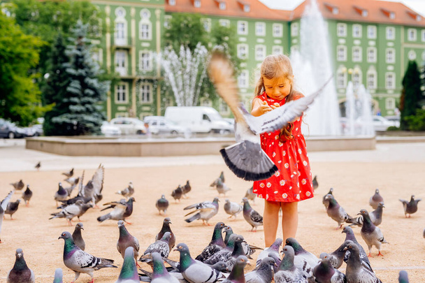 Junges kaukasisches weißes Mädchen füttert viele Tauben, einige fliegen herum oder setzen sich hin. Verschwommener Brunnen und grünes Gebäude im Hintergrund - Foto, Bild