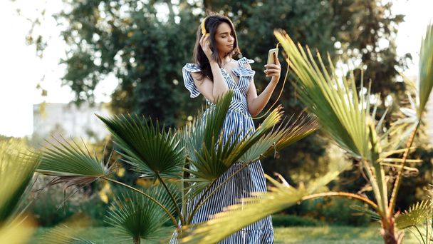 Attractive young woman in summer dress listening to music with headphones while holding mobile phone outdoor. - 写真・画像