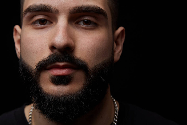 a close-up dramatic portrait of a young serious guy, musician, singer,rapper with a beard and arms crossed on his chest on a black isolated background. - Foto, Imagen