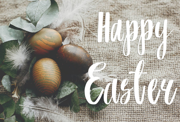 Happy Easter text. Easter greetings card. Stylish easter eggs with feathers on rustic table. Natural dyed green easter eggs with eucalyptus branch, spring flowers on rural textile - Φωτογραφία, εικόνα