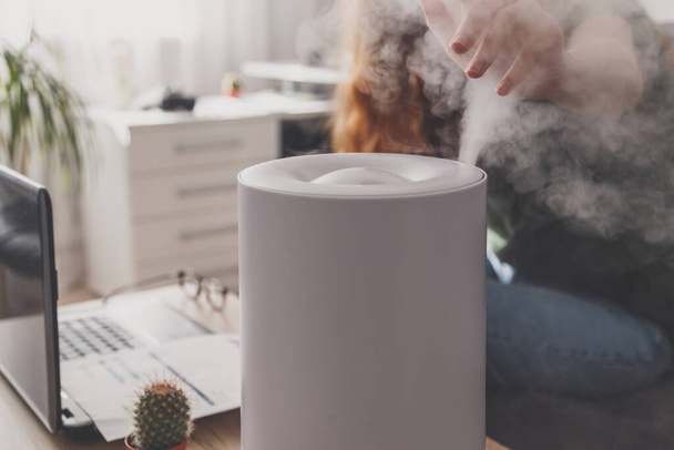 Woman freelancer uses a household humidifier in the workplace to maintain relative humidity and microclimate in the workplace of the home office with a laptop and documents. - 写真・画像