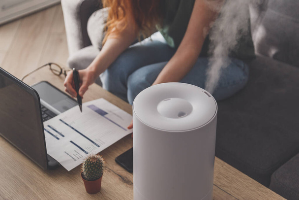 Woman freelancer uses a household humidifier in the workplace at home office with a laptop and documents. - Photo, image