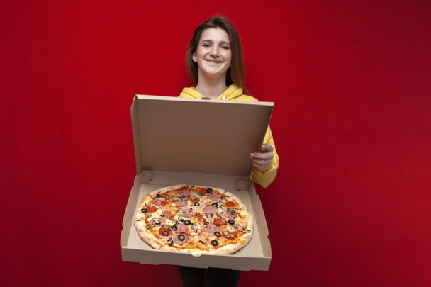 young beautiful cheerful girl holds a big pizza in a box on a red background, she opens and shows pizza, portrait of a pizza delivery girl, copy space - Valokuva, kuva