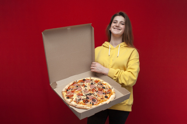 fiatal szép vidám lány tart egy nagy pizza egy dobozban egy piros háttér, kinyitja és mutatja pizza, portré egy pizza szállító lány, másolás helyet - Fotó, kép