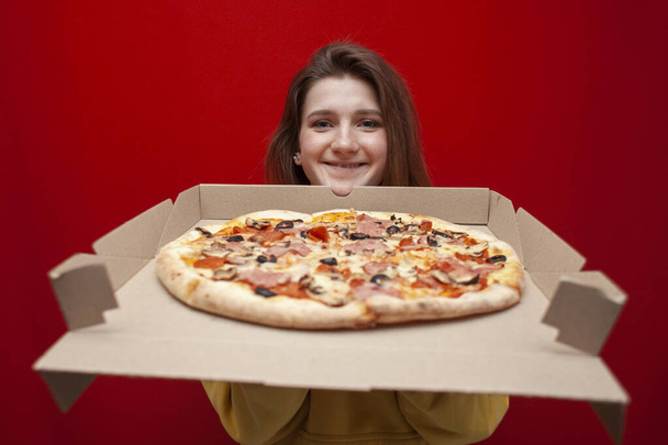 jong meisje student met een grote smakelijke pizza in een doos op een rode achtergrond, een hongerig meisje houdt fast food, kopieer ruimte - Foto, afbeelding