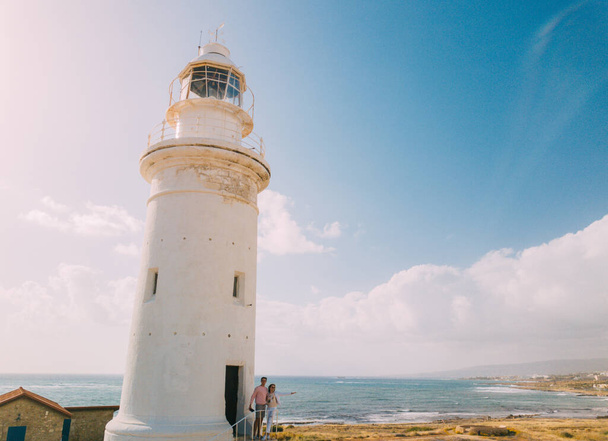 Beautiful young couple family bearded nordic man and trendy female in coat walking along sea shore with lighthouse hand in hand in. - Foto, Imagem