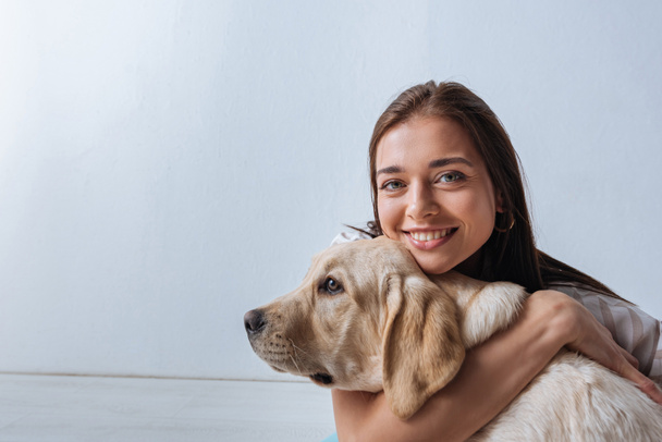 Beautiful woman smiling at camera while embracing golden retriever on grey background - 写真・画像