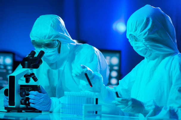 Scientists in protection suits and masks working in research lab using laboratory equipment: microscopes, test tubes. Medicine, coronavirus 2019-ncov infection and vaccine discovery concept. - Photo, image