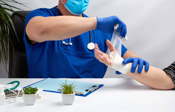 Άντρας γιατρός κάθεται σε ένα τραπέζι με μπλε στολή και γάντια λατέξ, ειδικός επιδέσμους το χέρι του με ένα λευκό επίδεσμο στον ασθενή, λαμβάνοντας ασθενείς στο γραφείο - Φωτογραφία, εικόνα