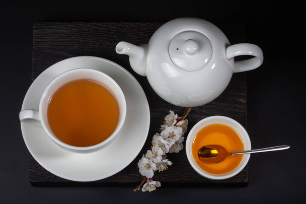 黒を基調とした白い磁器のお茶の絶景です。黒テーブルの上にお茶、ティーポット、蜂蜜の白い磁器カップ. - 写真・画像