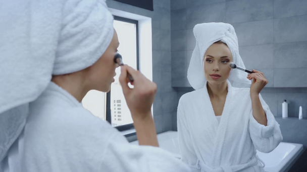 piękna młoda kobieta w szlafroku za pomocą pędzla kosmetycznego przed lustrem - Materiał filmowy, wideo