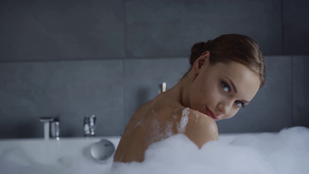 beautiful young woman washing shoulder and looking at camera in bubble bath - Footage, Video