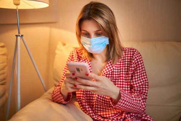 Young european woman wearing surgical face mask stay at home during coronavirus, covid-19 outbreak and looking news or application on smartphone - Фото, зображення