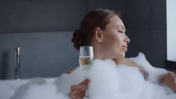 wunderschöne junge Frau schlürft Champagner und schaut in die Kamera im Schaumbad - Filmmaterial, Video