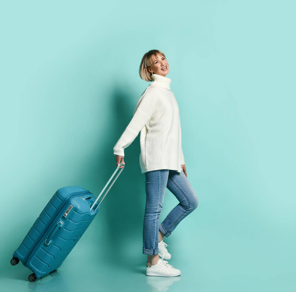 Blonde girl in white sweater, jeans and sneakers. She is smiling posing with suitcase luggage standing sideways on blue  - Foto, Imagem