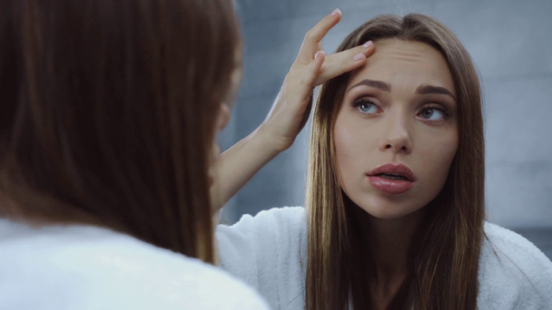 displeased young woman in bathrobe looking at face in mirror - Footage, Video