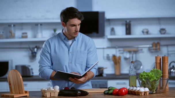 muž čtení kuchařky, zatímco stojí u stolu s čerstvými ingrediencemi - Záběry, video