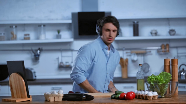 homem animado em fones de ouvido sem fio dançando perto da mesa com ingredientes frescos
 - Filmagem, Vídeo