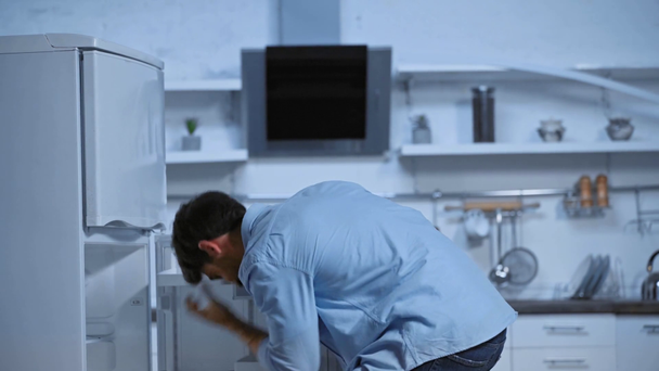 現代のキッチンで冷蔵庫を探している青いシャツの不快感の男 - 映像、動画