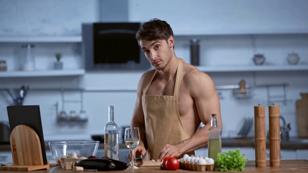 Hemdloser Mann in Schürze kocht in der Küche, blickt in die Kamera und lächelt - Filmmaterial, Video