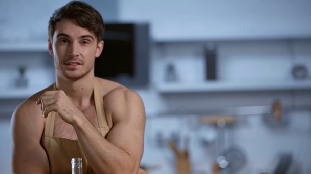shirtless man in apron holding hand near face and smiling at camera in kitchen - Footage, Video