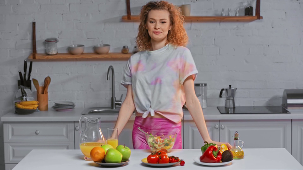Chica sonriente comiendo ensalada cerca de verduras frescas y frutas en la mesa
 - Imágenes, Vídeo
