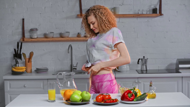 Uśmiechnięta dziewczyna mierzy talię przy warzywach, sałatce i soku pomarańczowym na stole - Materiał filmowy, wideo