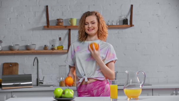 Mosolygó lány zsonglőrködik narancs közelében alma és narancslé az asztalon - Felvétel, videó