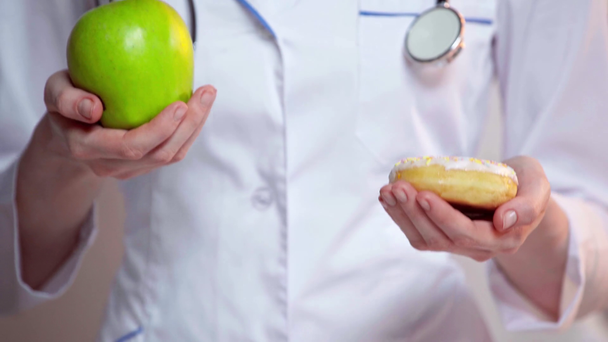 Oříznutý pohled na lékaře drží jablko a koblihu na šedém pozadí - Záběry, video