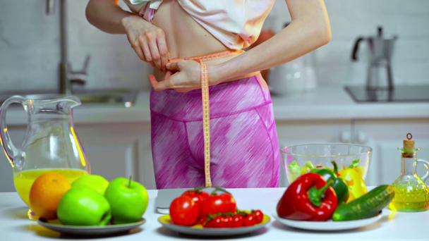 Vista cortada da mulher medindo a cintura perto de legumes e frutas na mesa
  - Filmagem, Vídeo