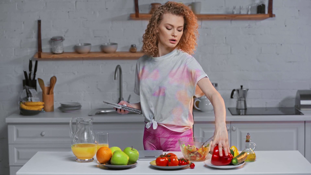 Žena s poznámkovým blokem o hmotnosti zeleniny v blízkosti salátu a pomerančového džusu na stole  - Záběry, video