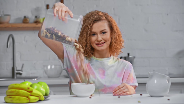 Mujer sonriente vertiendo cereales en un tazón en la cocina
  - Metraje, vídeo