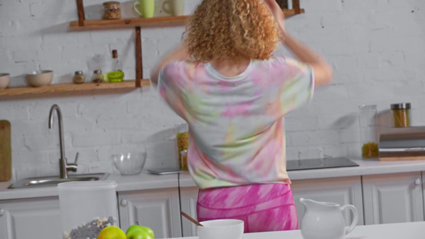 Dívka ve sluchátkách s chytrým telefonem tančí v blízkosti jablek a cereálií v kuchyni  - Záběry, video