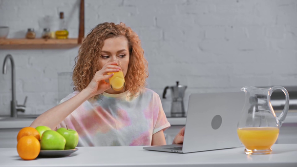 Lachende vrouw die laptop gebruikt en sinaasappelsap drinkt in de keuken  - Video