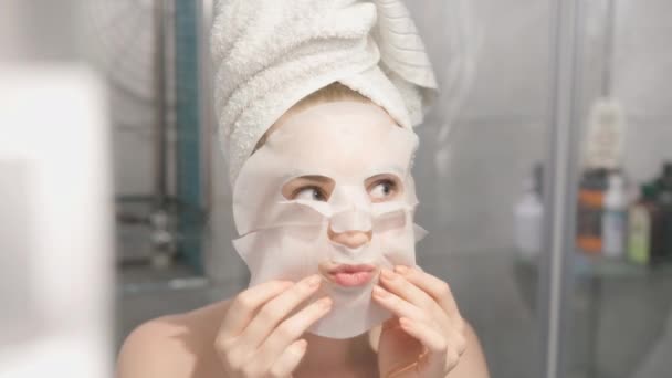 Woman applying cosmetic Japanese mask on her face and looking in the mirror looking at camera in the bathroom Blonde woman - Footage, Video
