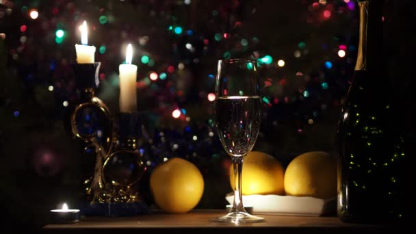 Holidays, Christmas, New year. Lights go up in a bottle that sits on a table with a glass, candles and oranges against a Christmas tree. Close up. - Filmati, video