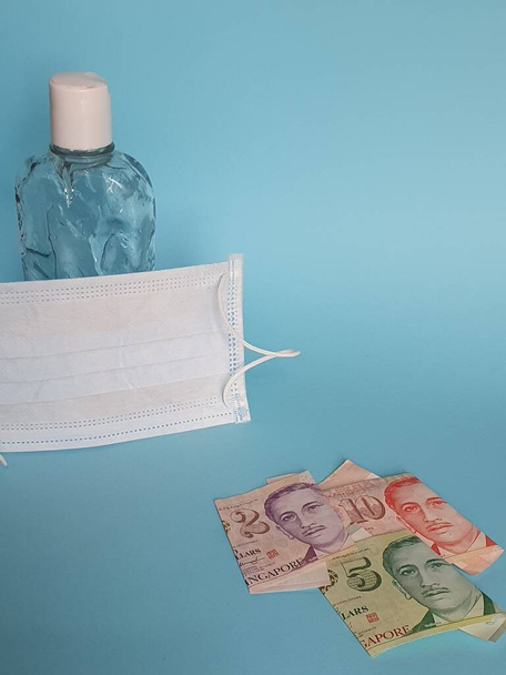 singaporean bankbiljetten, gezichtsmasker, fles met gel alcohol en blauwe achtergrond - Foto, afbeelding