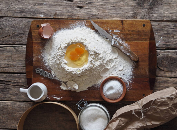 Pastry concept.On the kitchen cutting Board, a broken egg in flour with ingredients for making dough and kitchenware. Home cooking.Ancient wooden background.The view from the top.Space for text. - Fotó, kép
