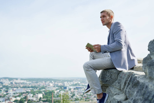 businessman with a tablet in his hands sits on a rock against the background of the city and looks into the distance - Fotó, kép