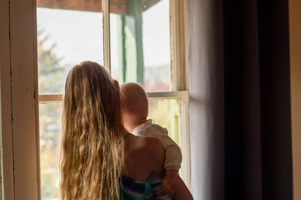 Quédate en casa: una chica rubia sostiene a su hermano pequeño cerca de la ventana. Concepto de autoaislamiento coronavirus
 - Foto, Imagen